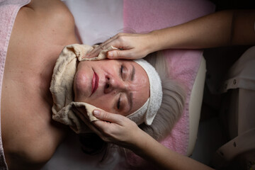 Close up of a mature woman receiving a relaxing facial massage from a professional female therapist in a modern spa. The client lies on a treatment bed with a towel and headband, enjoying skincare