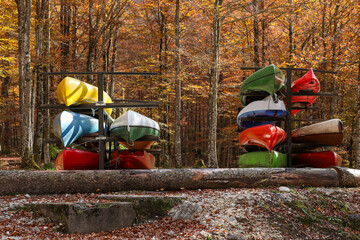 Bright kayaks and canoes are stacked on metal racks in an autumn forest. The vivid boats contrast with golden trees, capturing a peaceful fall outdoor scene with sunlight filtering through the leaves.