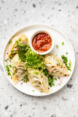 Steamed dumplings (momos) served on a white plate with a dipping sauce, garnished with fresh herbs. The dish is presented on a minimalist gray background, top view.