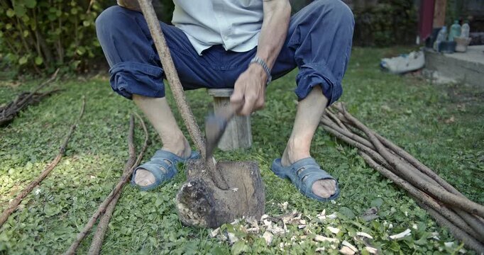 An elderly man diligently uses an axe to debark wood branches, shaping them with traditional craftsmanship in a rustic outdoor environment.