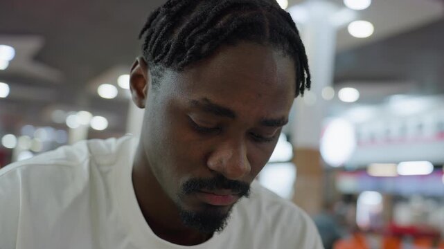Young african man with braided hair and goatee wearing white shirt sits indoors looking around with contemplative expression, surrounded by blurred bright lights and background