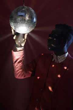 Fototapeta Portrait of fashionable African American guy in sunglasses and suit twisting shiny disco ball while standing in the dark studio