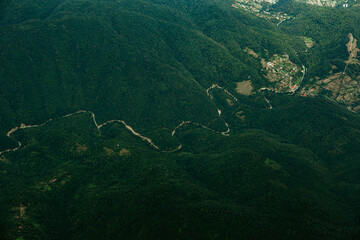 Aerial view of a winding road cutting through lush green mountain hills. Scenic landscape showing natural curves, dense forest, and elevation.