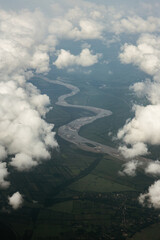 River Flowing Between Clouds Seen from Airplane. Aerial view of a winding river flowing through green fields and farmland, partially covered by soft white clouds.