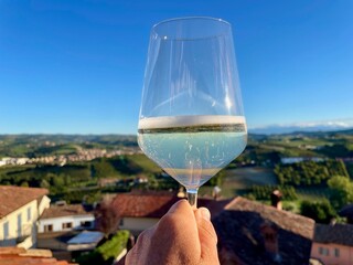 A hand elegantly raising a glass of sparkling wine before a panorama of sunlit vineyards, Roddi, Piedmont.