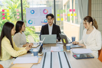Group of Asian business people presenting data to colleagues in conference room. Business people in suits holding pens pointing at graph documents on table to present company data.