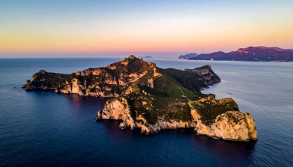 An aerial view of a rugged, elongated island at sunset, with cliffs and vegetation. Gentle waves lap against the shore. Another smaller landmass in the distance