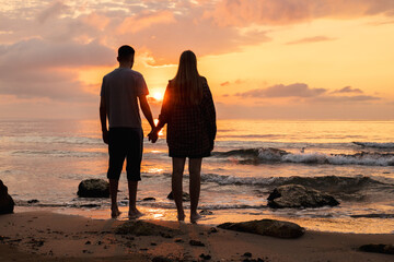 Couple holding hands and watching the sunrise over the sea, standing barefoot on a sandy beach with gentle waves and warm morning light