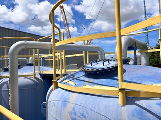 Large industrial water treatment facility with blue filtration tanks, yellow railings, and complex piping under a bright, cloudy sky.