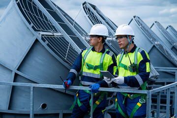 Two men in safety gear are standing on a railing looking at a large fan