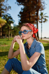lifestyle woman wearing modern boho-western denim outfit and blue sunglasses sitting outdoors with thoughtful expression in warm film color lighting