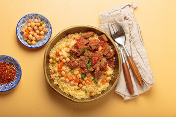 Traditional dish of Maghreb cuisine, a Moroccan couscous with lamb or beef, vegetables, chickpeas, and peas in a rustic bowl with cutlery on yellow background