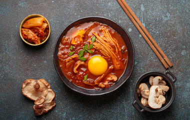 Top view of spicy kimchi jjigae soup in a black bowl with egg yolk, enoki, shiitake, tofu and green onions, with chopsticks and cooking ingredients, cuisine of Korea