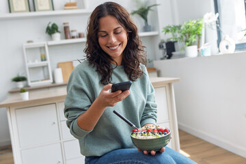 Pretty woman eating a healthy fruit bowl while using her smartphone in the kitchen at home