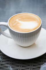 A freshly made cappuccino with delicate milk froth sits on a white saucer atop a woven café table.