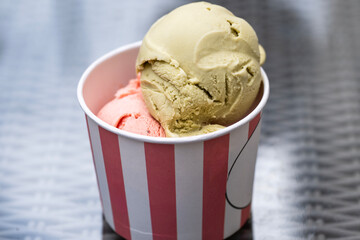 Two scoops of colorful ice cream sit in a striped paper cup on a metallic textured surface.