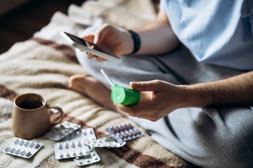 Obraz premium Man on bed holding smartphone and throat spray surrounded by medicine at home.
