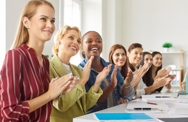 Diverse group of women sitting at desk, clapping hands and smiling enthusiastically. Happy multiracial female team attending event, training or presentation, showing support and appreciation to coach.