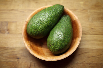 Two fresh, ripe green avocados  placed in a rustic wooden bowl on a natural wood background, highlighting healthy eating, cooking, and natural food ingredients.