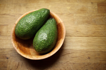Two fresh, ripe green avocados  placed in a rustic wooden bowl on a natural wood background, highlighting healthy eating, cooking, and natural food ingredients.