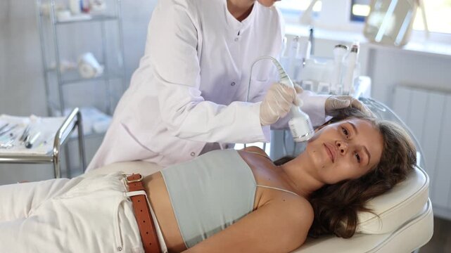 Positive young girl undergoing facial treatment using cold hammer helping to shrink pores and relieve skin inflammation in aesthetic medicine clinic. Modern hardware cosmetology. High quality 4k