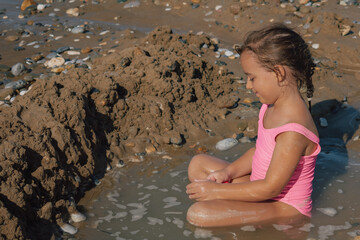 A happy little girl smiling and enjoying playing in the wet sand and shallow water on a pebble beach, sitting next to her handmade sand fort on a sunny day