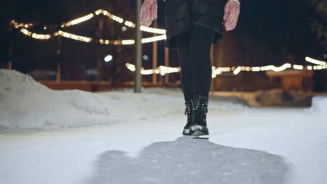 Person wearing winter boots and warm clothing walking on snowy path at night with glowing festive lights in background, shadow stretching on snow, capturing cozy atmosphere of cold evening stroll