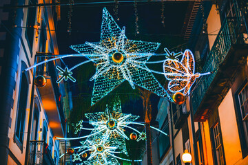 Street at night decorated with festive Christmas lights. Large star-shaped and butterfly-shaped illuminations hang above the street, brightening the dark buildings