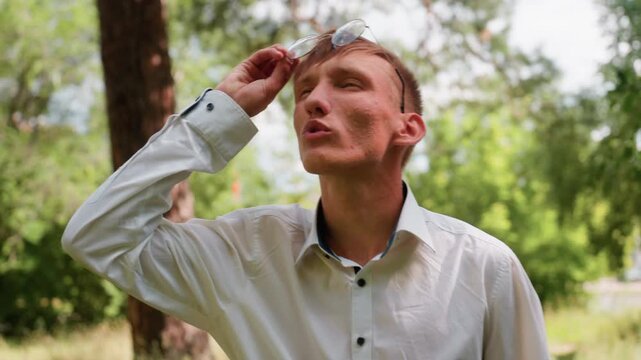 Close view of young man in white shirt raising glasses while whistling with mouth and observing trees in natural forest setting, surrounded by green foliage, expressing attention