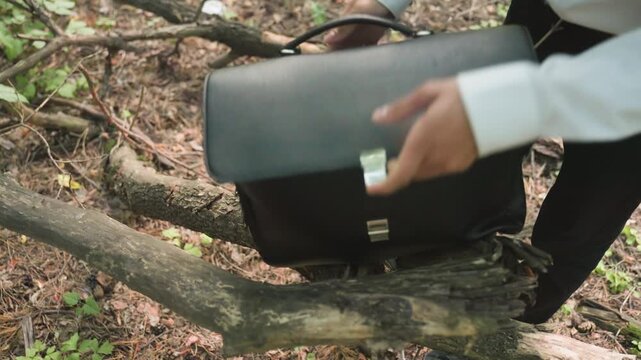 Close-up view of ecologist in white shirt placing white coat inside black bag resting on fallen tree in forest, surrounded by greenery and natural sunlight creating woodland atmosphere