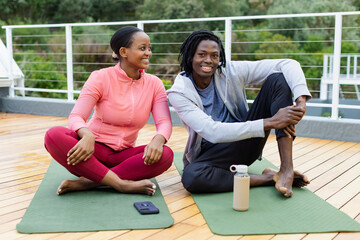 African American couple sitting on green mats on deck exchanging smiles with smartphone and bottle
