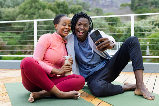 African American couple taking phone selfie sitting on yoga mats on terrace holding water bottle - Powered by Adobe