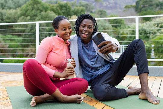 African American couple sitting on yoga mats on wooden deck holding water bottle and smartphone