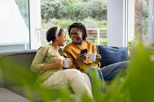 African American couple smiling and chatting on sofa in living room holding mugs and smartphone