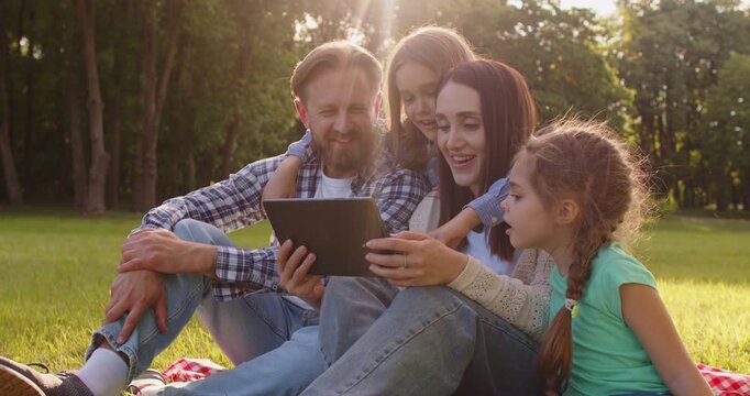 Father, mother, daughter and son spending leisure time together in park or at field, using digital tablet for watching film online or scrolling internet, being always in touch. Family leisure 