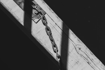 Rusty chain attached to weathered wooden beam over dark water. Old metal chain and shadow detail on sunlit dock surface.