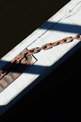 Rusty chain attached to weathered wooden beam over dark water. Old metal chain and shadow detail on sunlit dock surface.