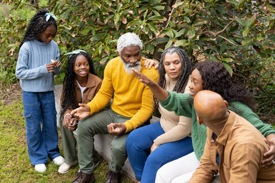 Diverse multigenerational family chatting around stone bench in park wearing sweaters, sturdy shoes
