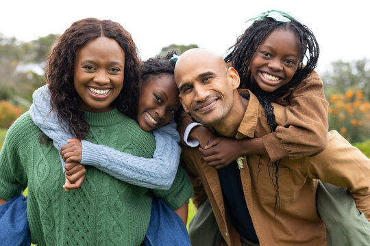 African American family carrying daughters piggyback while smiling in orchard with orange blossoms
