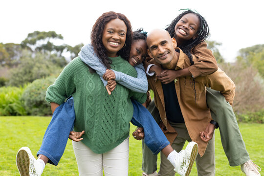 Diverse family giving piggyback rides on grassy lawn in public park under cloudy sky, smiling