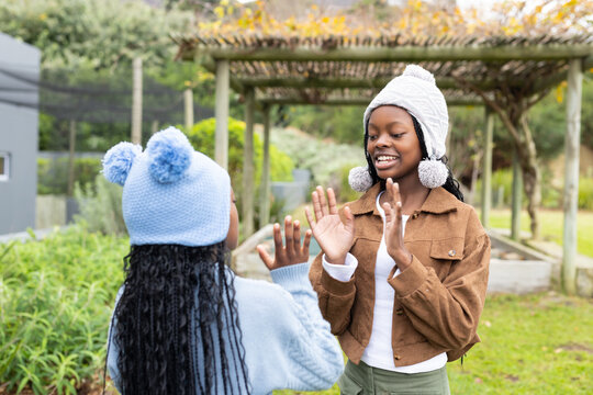 African American schoolgirls wearing pom-pom hats playing clapping game beneath pergola in garden