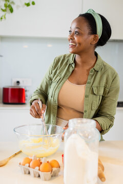 African American woman smiling while whisking batter at kitchen island with eggs and flour jar