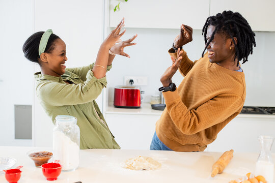 African American couple laughing and raising hands playfully in kitchen with flour jar, rolling pin