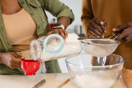 Black couple in kitchen pouring jar of flour into cup and sifting through sieve over bowl - Powered by Adobe