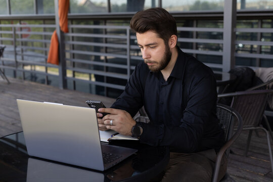 The man is looking at his phone while sitting at a table with a laptop
