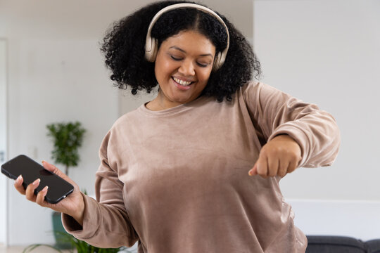African American mid adult woman dancing and smiling in living room with smartphone and headphones