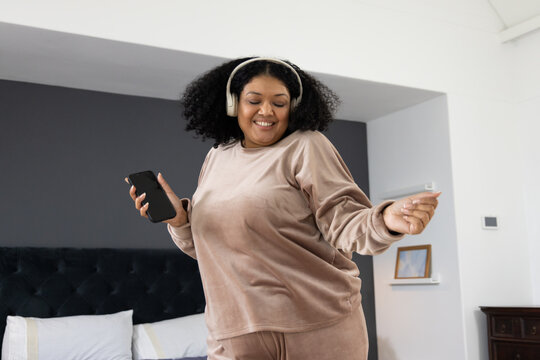 African American woman dancing in bedroom holding smartphone wearing headphones by headboard - Powered by Adobe