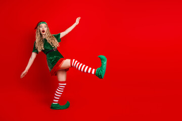 Festive young woman in a cheerful elf costume striking a playful pose on a vibrant red background...