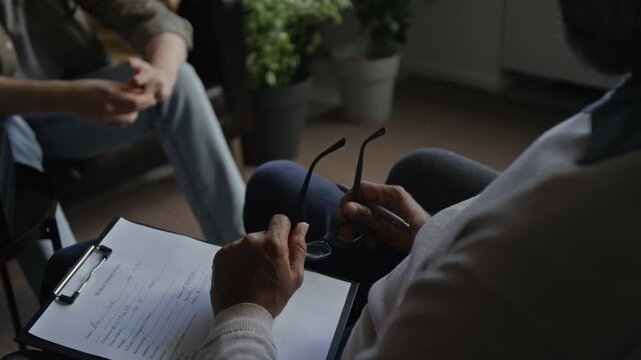 Over shoulder closeup of male therapist holding glasses and clipboard while listening to unrecognizable couple in therapy session, copy space