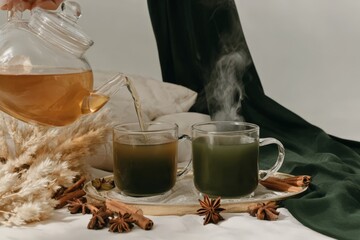 Elegant tea setup with glass teapot and mugs surrounded by spices and dried grass